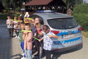 Dzieci wraz z policjantką przy radiowozie, prezentują odblaskowe opaski