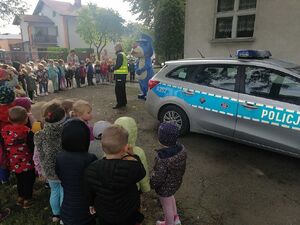 Policjant stoi przed radiowozem i prowadzi zajęcia profilaktyczne