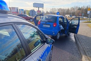 Na zdjęciu widać radiowóz, przed który stoi zatrzymany w wyniku pościgu samochód. W tle widać policjanta, który kieruje ruchem.