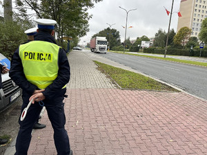 Na zdjęciu widać policjantów ruchu drogowego, a w tle radiowóz i jezdnia po której porusza się samochód ciężarowy.