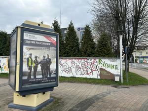 Na zdjęciu widać ekspozytor ogłoszeniowy, w którym znajduje się reklama zatrudnienia w Policji na tle ogrodzenia i budynku żorskiej Policji.