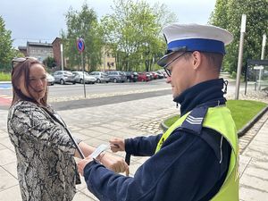 Na zdjęciu widać policjanta w białej czapce i odblaskowej kamizelce, który przekazuje odblaskową listwę kobiecie.