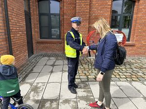 Na zdjęciu widać policjanta w białej czapce i odblaskowej kamizelce, który przekazuje odblaskową listwę kobiecie z dzieckiem.