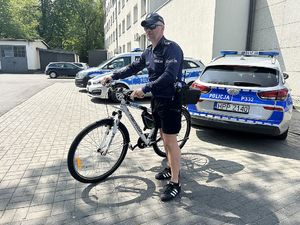 Na zdjęciu widać policjanta przy służbowym rowerze, a tle znajduje się teren żorskiej Policji i radiowozy.