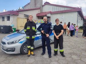 Na zdjęciu widać policjanta przed radiowozem, a obok stoją strażacy.