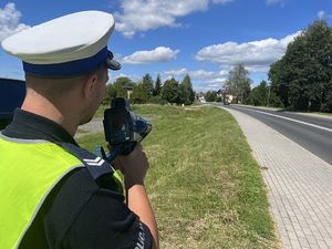 Na zdjęciu widać policjanta ubranego w kamizelkę odblaskową, który mierzy prędkość nadjeżdżających pojazdów.