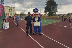 Na zdjęciu widać policjanta oraz Sznupka, a w tle znajduje się boisko sportowe.