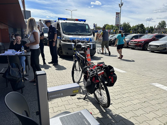 Na zdjęciu widać policjanta, policyjny radiowóz, a na pierwszym planie urządzenie do znakowania rowerów oraz rower.