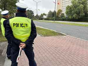 Na zdjęciu widać policjantów ubranych w kamizelki odblaskowe, którzy stoją przy drodze. Jeden z policjantów w dłoni trzyma tarczkę do zatrzymywania pojazdów.
