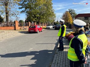 Na zdjęciu widać policjantów ruchu drogowego ubranych w kamizelki odblaskowe oraz białe czapki. Mundurowi stoją w rejonie cmentarza.