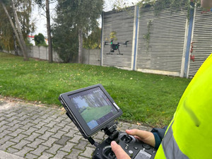 Na zdjęciu widać policjanta ubranego w odblaskową kamizelkę trzymającego w dłoniach urządzenie do sterowania dronem.