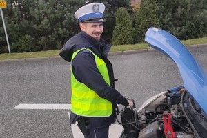 Na zdjęciu widać policjanta, którzy stoi przed niebieskim samochodem. W dłoniach trzyma tzw. &quot;starter&quot; służący do uruchamiania pojazdu, któremu rozładował się akumulator.