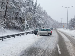 Na zdjęciu na pierwszym planie widać mocno uszkodzony w przedniej części srebrny samochód. W tle widać drogę i las.