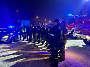 Na zdjęciu widać policjantów i strażaków oddających hołd poległym policjantom. W tle widać wozy strażackie i policyjny radiowóz.