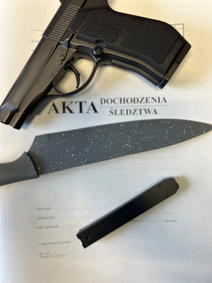 Na zdjęciu widać pistolet koloru czarnego oraz nóż kuchenny ułożone na teczce z napisem akta dochodzenia/śledztwa.
