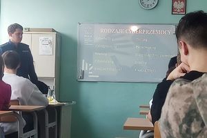 Na zdjęciu widać policjanta oraz uczniów, a w tle wyświetlana jest prezentacja dotycząca rodzajów cyberprzemocy.