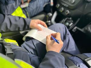 Na zdjęciu widać policjanta siedzącego w radiowozie i wypisującego mandat karny.