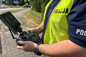 Na zdjęciu widać umundurowanego policjanta trzymającego w dłoniach urządzenie do sterowanie dronem. W tle widać oznakowane przejście dla pieszych i przejeżdżające pojazdy.