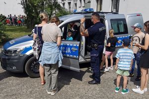 Na zdjęciu widać policjanta przy radiowozie, wewnątrz &quot;za kierownicą&quot; siedzi chłopiec, wokół uczestnicy festynu.