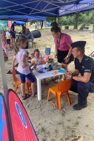 Na zdjęciu widać policjanta, który przykuca przy stoliku, dzieci wykonują zadania.