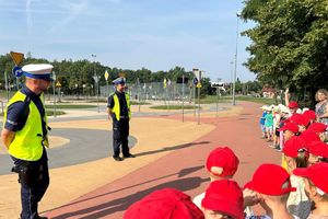 Na zdjęciu widać policjantów, wychowawców i przedszkolaków na terenie miasteczka ruchu drogowego. Dzieci ubrane są w kolorowe czapki z daszkiem.