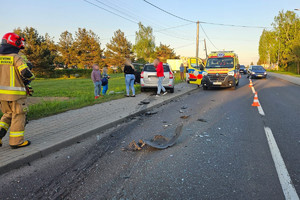 Na zdjęciu widać drogę, pozostałe na jezdni elementy uszkodzonego pojazdu, karetkę pogotowia, strażaka oraz uczestników zdarzenia.