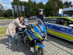 Na zdjęciu widać motocykl policyjny. Na motocyklu siedzi mały chłopiec, obok motocykla po lewej stronie stoi kobieta, po prawej stoi policjant. W tle widać radiowóz polskiej Policji.