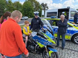 Na zdjęciu widać chłopca siedzącego na motocyklu policyjnym. Po lewej stronie widać kobietę oraz mężczyznę. Po prawej widać policjantów oraz radiowozy polskiej Policji.