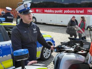 Na zdjęciu widać policjanta opierającego się o motocykl policyjny. W tle widać ambulans krwiodawstwa, uczestników pikniku i radiowóz polskiej policji.