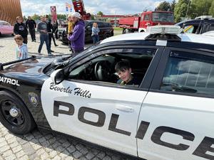 Na zdjęciu widać amerykański radiowóz oraz siedzącego w nim chłopca. Radiowóz jest czarno biały z napisem POLICE. W tle widać uczestników pikniku oraz amerykański radiowóz oraz stary wóz strażacki.