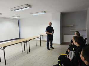 Na zdjęciu widać policjanta oraz uczestników spotkania zgromadzonych w sali odpraw.