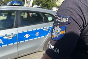Na zdjęciu widać policjanta, a w tle radiowóz. Na ramieniu ma odznaczenie o treści &quot;Policja, Pomagamy i chronimy&quot;.