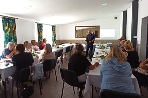 Na zdjęciu widać policjanta oraz uczestników debaty w sali ogrodów działkowych &quot;Aster&quot;.