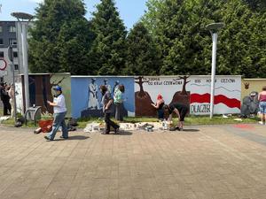 Na zdjęciu widać galerię murali podczas ich tworzenia, a także uczniów i zgromadzonych podczas akcji pod nazwą &quot;Plenerowa lekcja patriotyzmu&quot;.