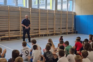 Na zdjęciu widać policjanta oraz zgromadzonych w sali gimnastycznej uczniów.