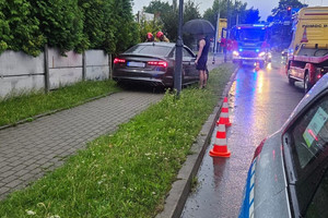 Na zdjęciu widać ciemny samochód, pojazdy służb ratunkowych i miejsce zdarzenia drogowego.