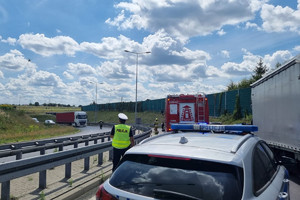 Na zdjęciu widać uszkodzony motocykl, radiowóz, policjanta oraz śmigłowiec LPR, w tle widać fragment autostrady.