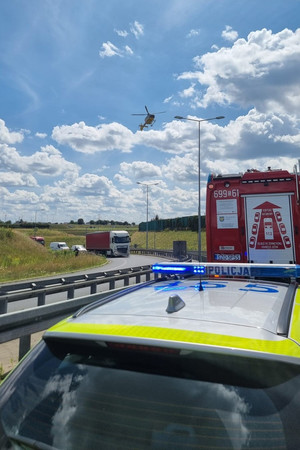 Na zdjęciu widać uszkodzony motocykl, radiowóz, policjanta oraz śmigłowiec LPR, w tle widać fragment autostrady.