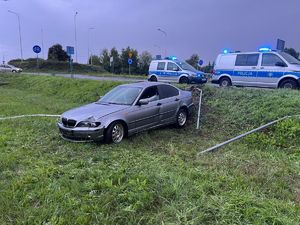 Na miejscu widać uszkodzony szary samochód, drogę, uszkodzone znaki oraz radiowozy policyjne.