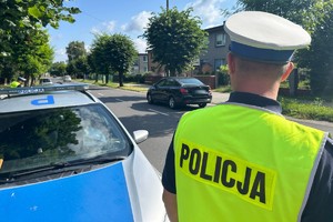 Na zdjęciu widać policjanta ubranego w białą czapkę, a obok policyjny radiowóz.