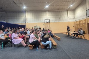 Na zdjęciu widać rodziców uczniów zgromadzonych w sali oraz policjanta i przedstawiciela szkoły.