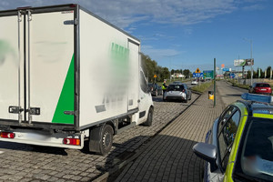 Na zdjęciu widać policjanta, radiowóz oraz kontrolowany samochód. W tle widać zabudowania i drogę.