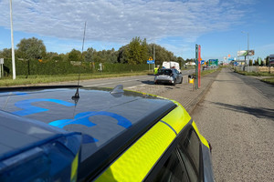 Na zdjęciu widać policjanta, radiowóz oraz kontrolowany samochód. W tle widać zabudowania i drogę.