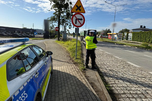 Na zdjęciu widać policjanta, radiowóz oraz kontrolowany samochód. W tle widać zabudowania i drogę.