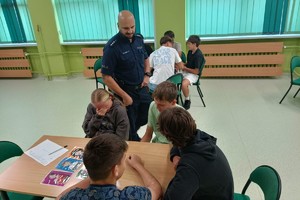 Na zdjęciu widać policjanta w sali lekcyjnej oraz uczniów siedzących w ławkach.