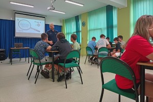 Na zdjęciu widać policjanta w sali lekcyjnej oraz uczniów siedzących w ławkach.