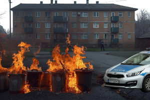 Grafika przedstawia palące się kosze na śmieci, w tle widać budynek i mężczyznę, a na pierwszym planie policyjny radiowóz.