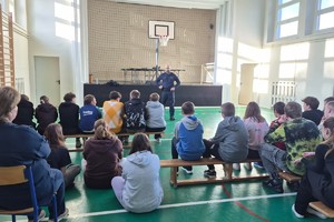Na zdjęciu widać policjanta oraz uczniów zgromadzonych w sali gimnastycznej.