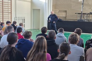 Na zdjęciu widać policjanta oraz uczniów zgromadzonych w sali gimnastycznej.