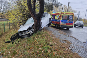Na zdjęciu widać uszkodzony srebrny samochód, miejsce zdarzenia drogowego, radiowóz oraz ambulans pogotowia.